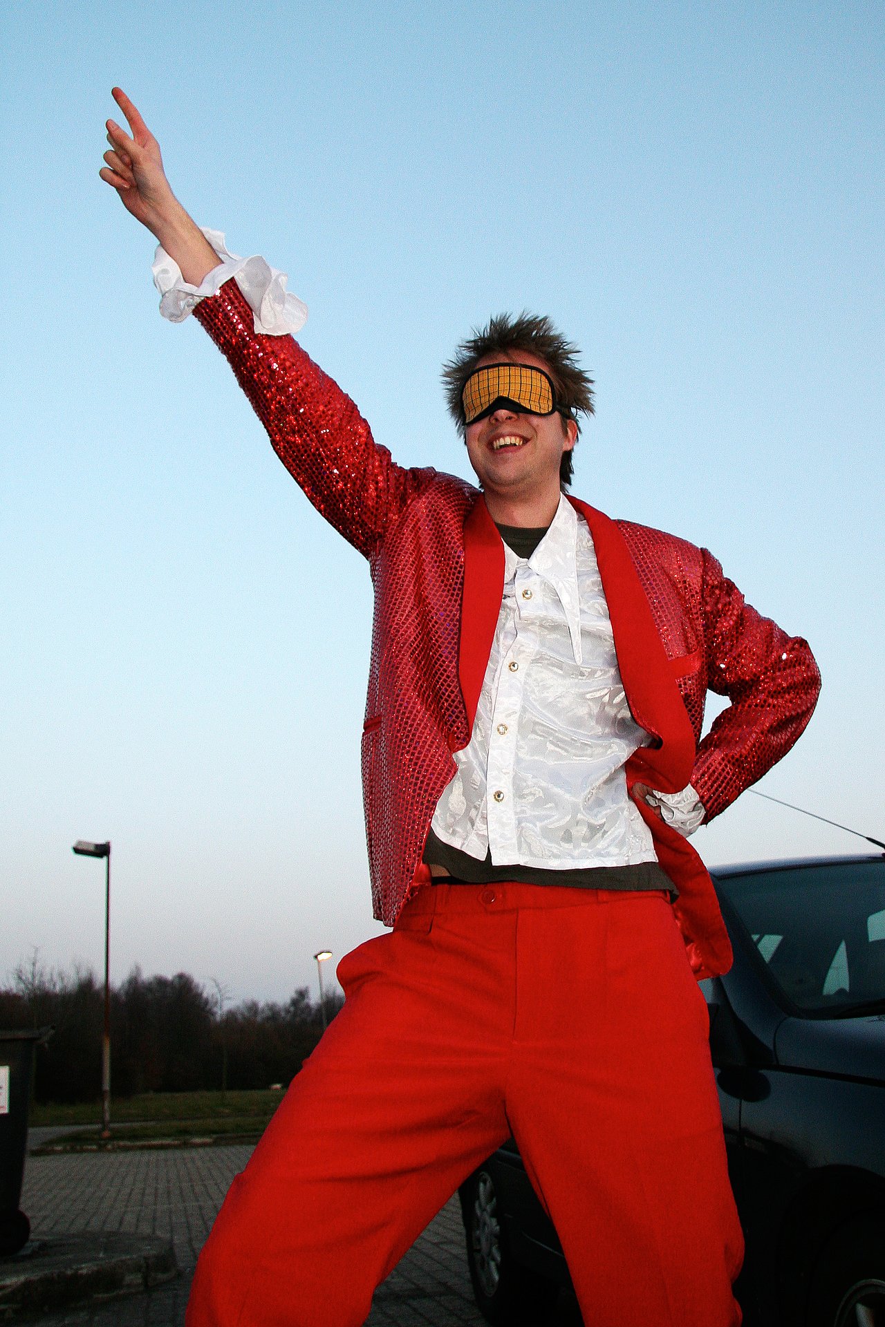 A blindfolded man in a red sequined outfit raises one arm and smiles during his bachelor party.