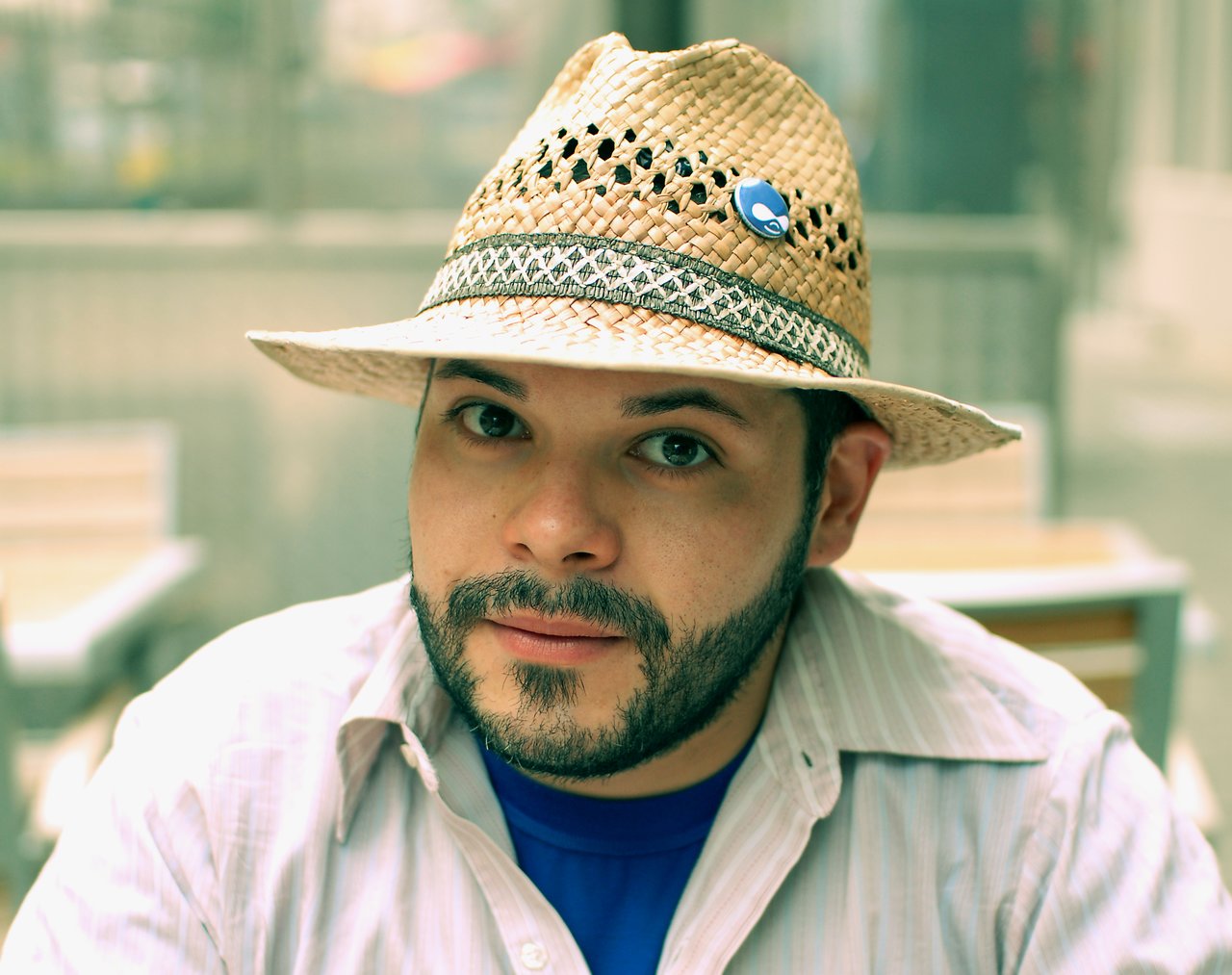 A bearded man wearing a straw hat and a button-up shirt sits on a terrace, looking directly at the camera.