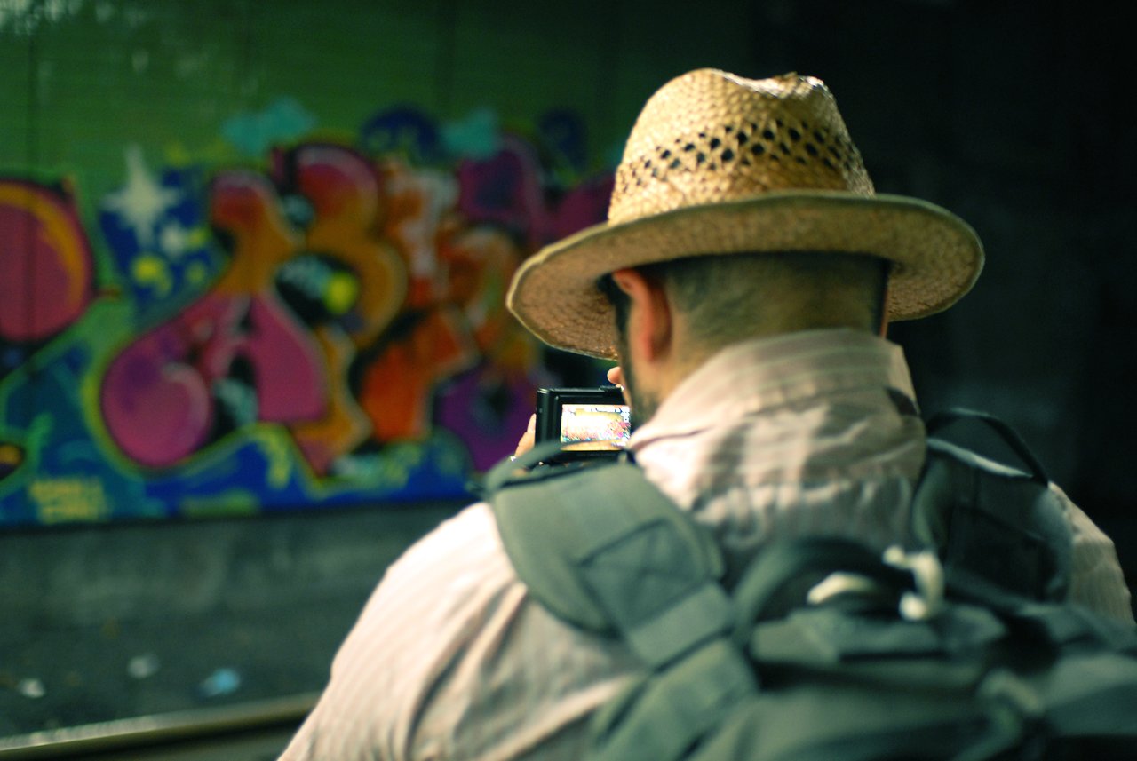 A person wearing a straw hat and backpack takes a photo of colorful graffiti in a subway station.