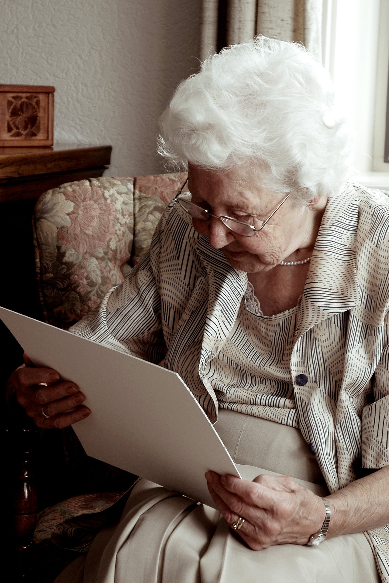 An elderly woman with white hair and glasses looks at a wedding photo album while sitting in a chair.