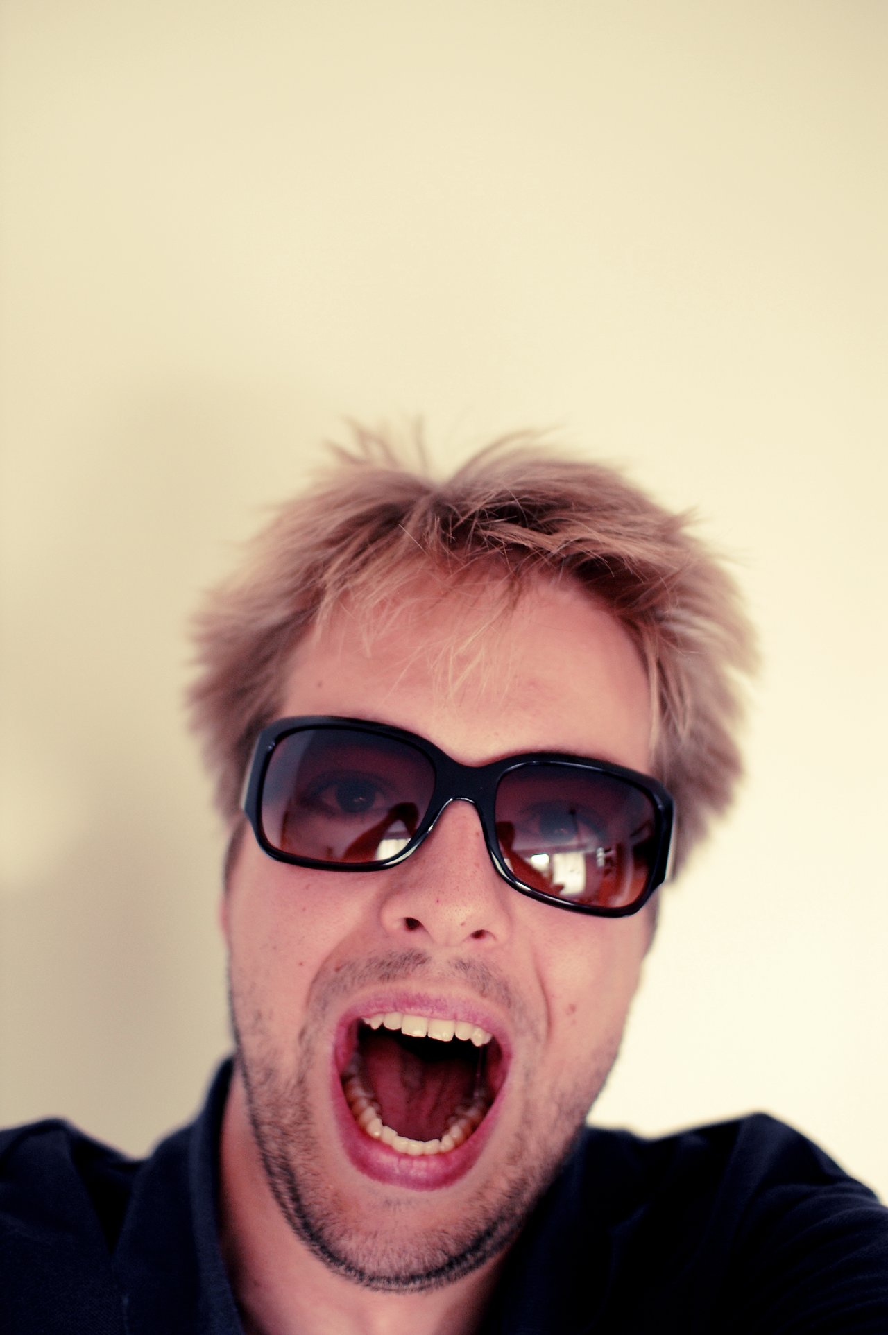 A person with sunglasses and messy hair opens their mouth wide, possibly shouting or reacting excitedly.