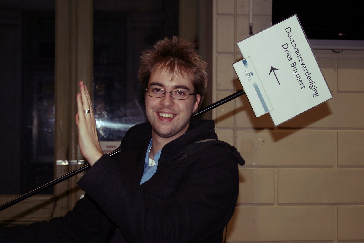 A smiling person in a dark coat holds a sign pointing to a PhD defense location while waving.