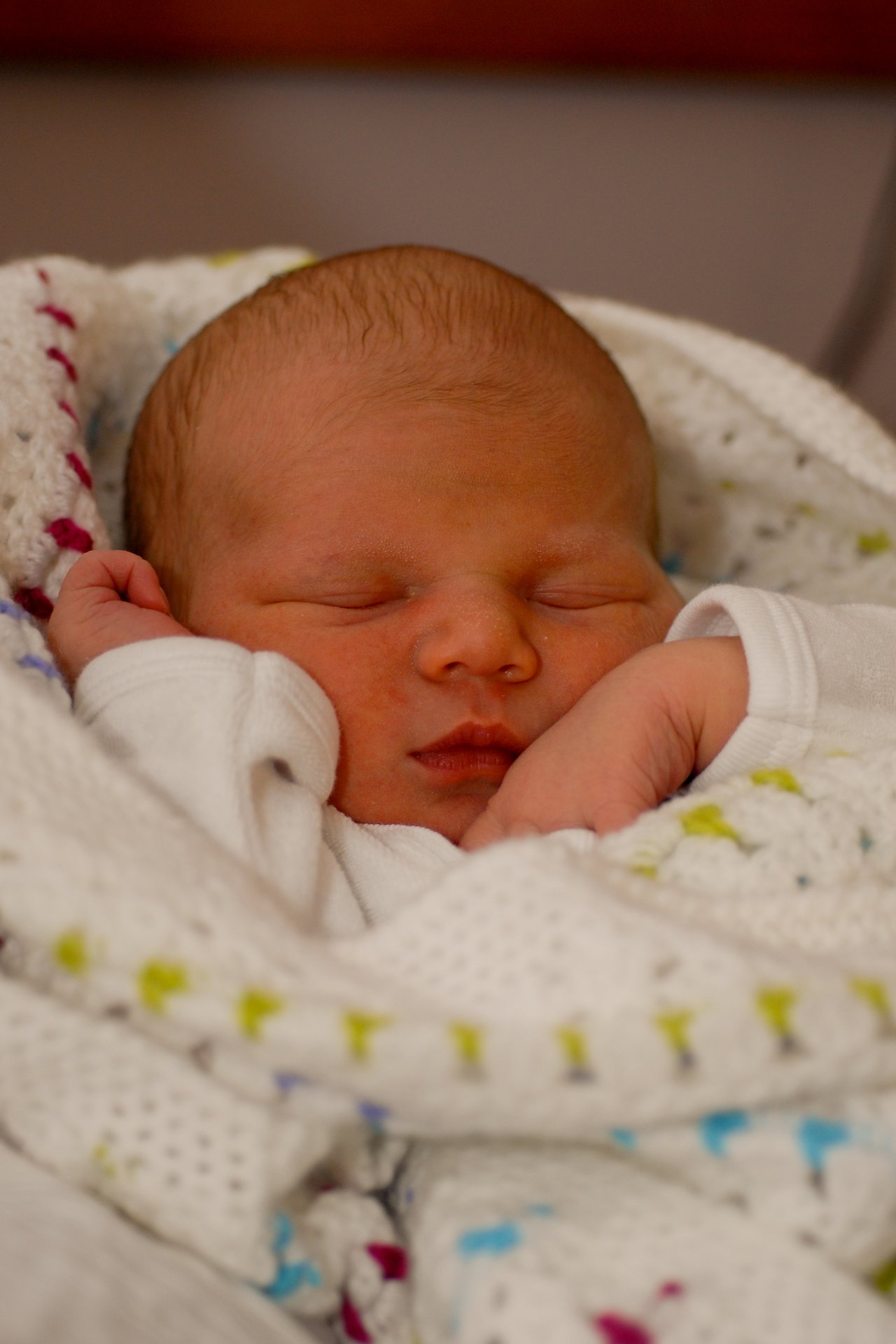 A newborn baby sleeps peacefully, wrapped in a soft, white knitted blanket with colorful accents.