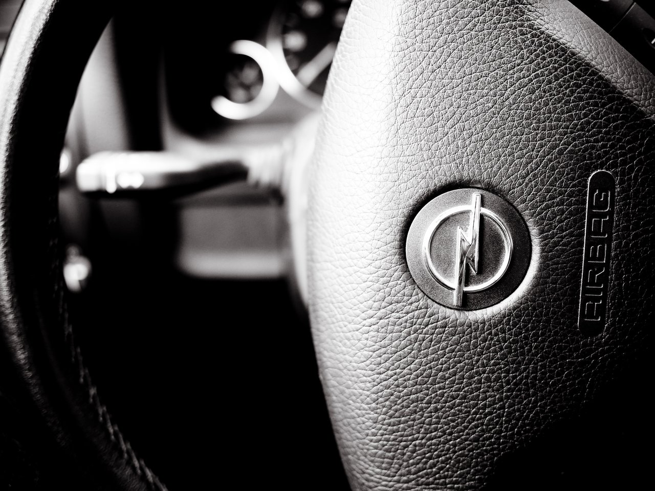 Close-up of an Opel steering wheel, showing the logo and textured surface, with dashboard controls slightly visible in the background.