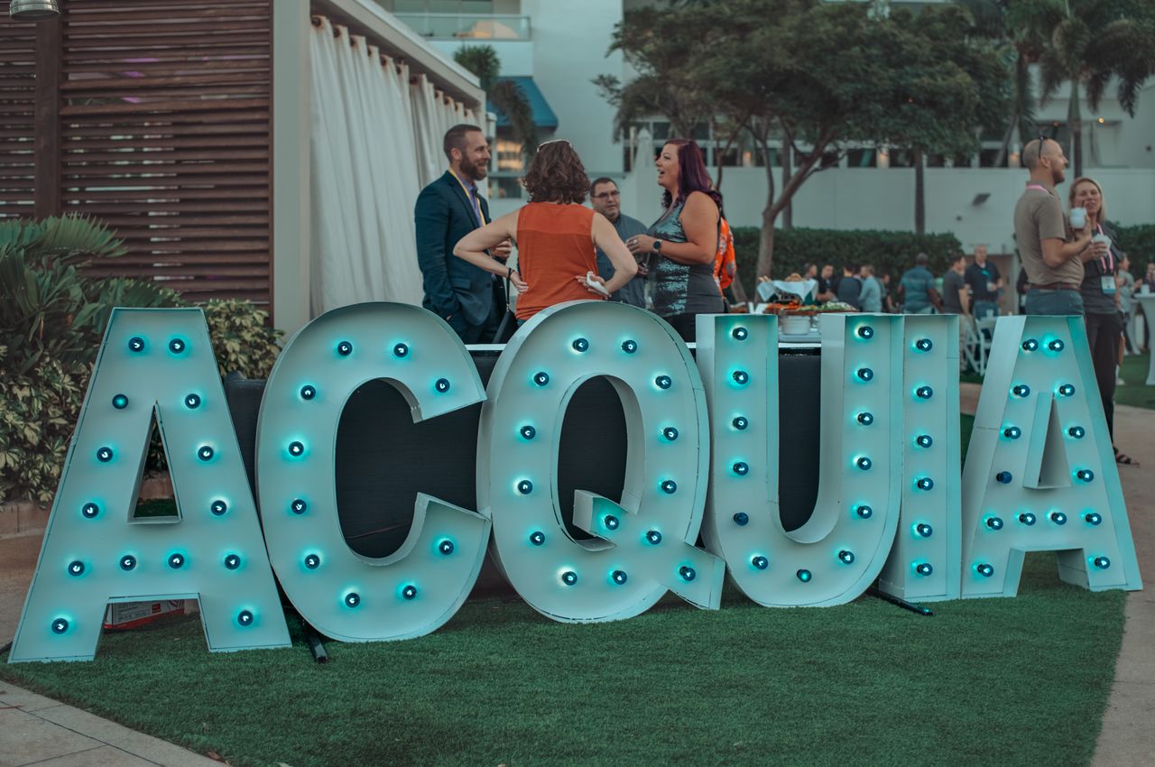 A large sign that spells out 'Acquia' in individually lit letters. Behind the sign, people are engaged in a lively conversation.