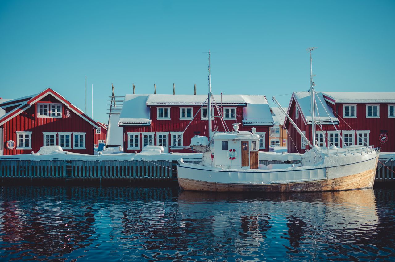 A old, snow-covered fishing boat docked in front of red buildings that serve as homes for fishermen.