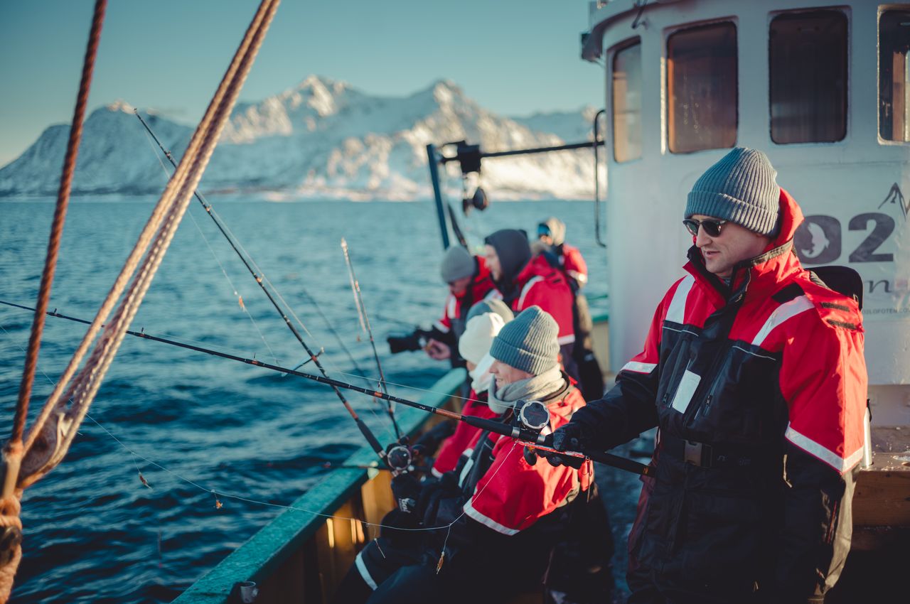 People with fishing rods on a boat, bundled up to stay warm in the cold arctic weather. Snow-covered mountains provide a beautiful backdrop.