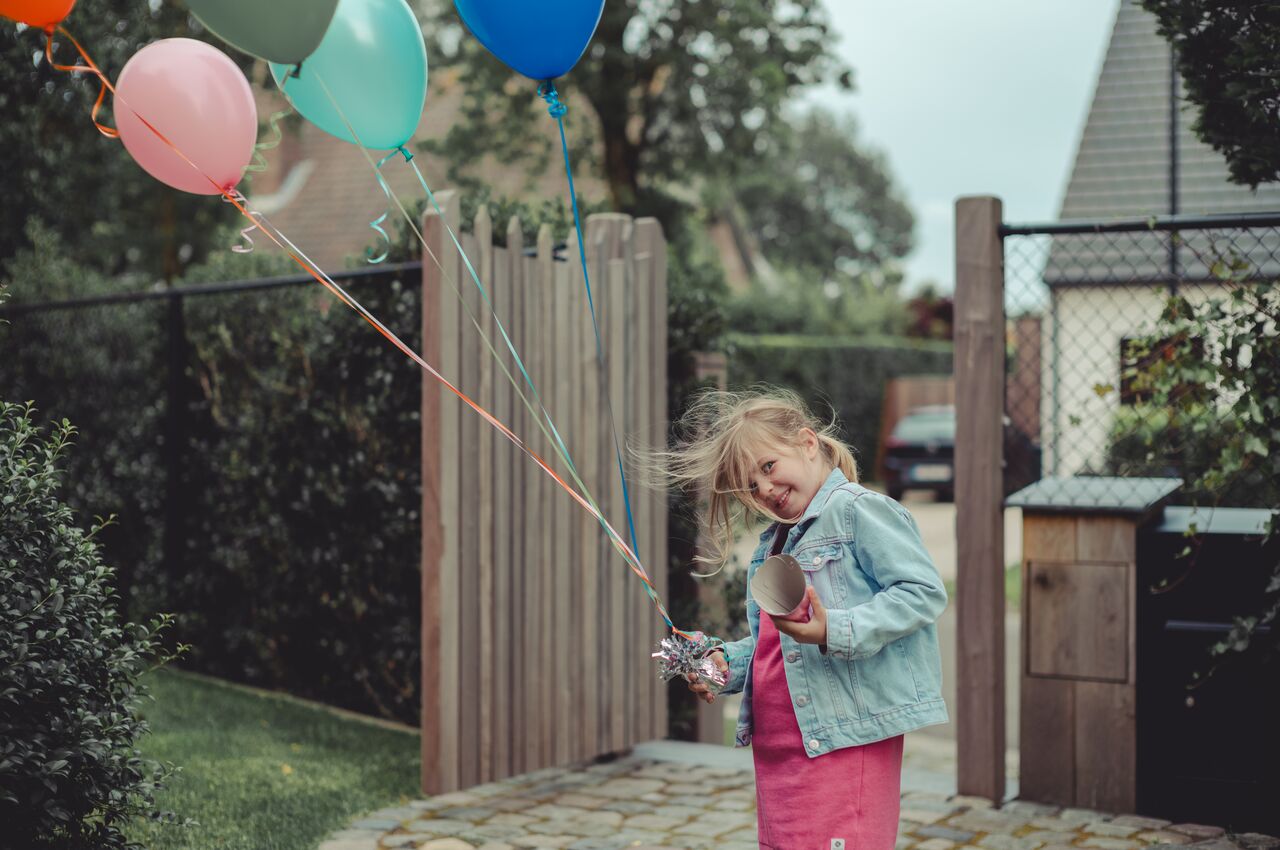 A smiling girl in a denim jacket and pink dress holds a bunch of colorful balloons by a wooden garden gate, her hair blowing in the wind.