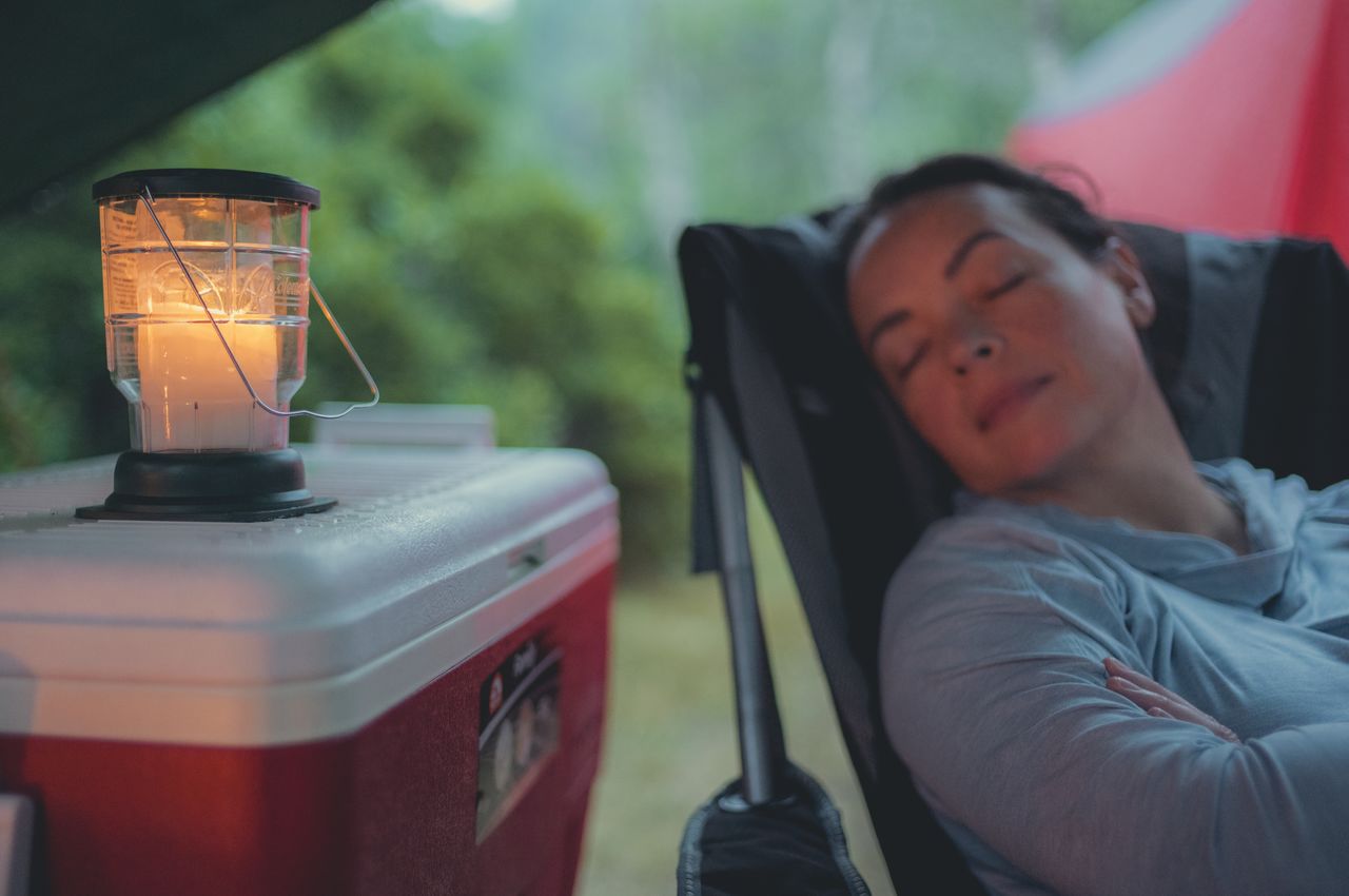 A candle on top a cooler, gently casting a warm glow on a person peacefully asleep in a camping chair.