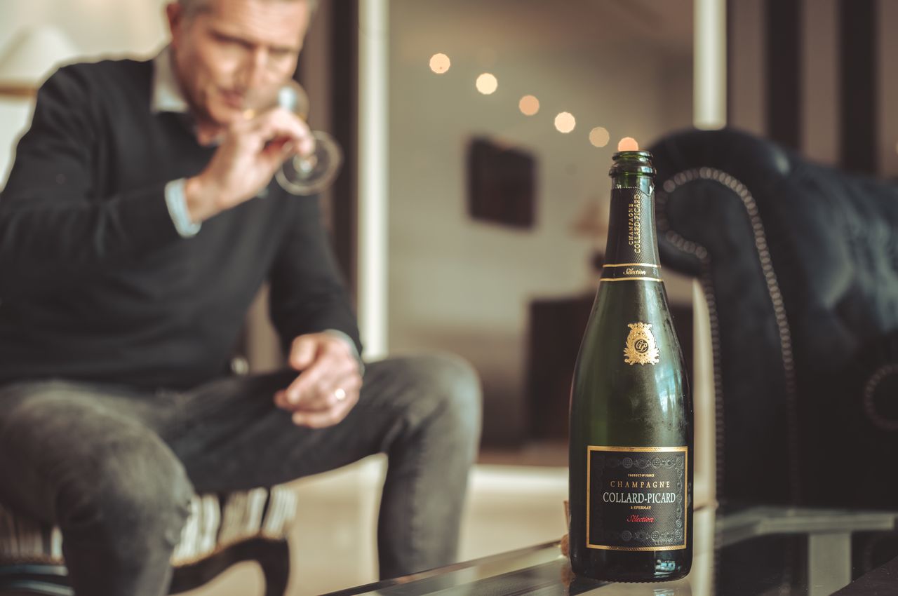 An open bottle of Collard-Picard champagne in focus, with a man drinking champagne in the background.
