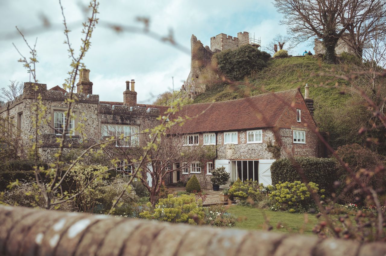 A quaint old cottage nestled at the base of a hill, surrounded by trees and flowers. An old castle sits atop the hill.
