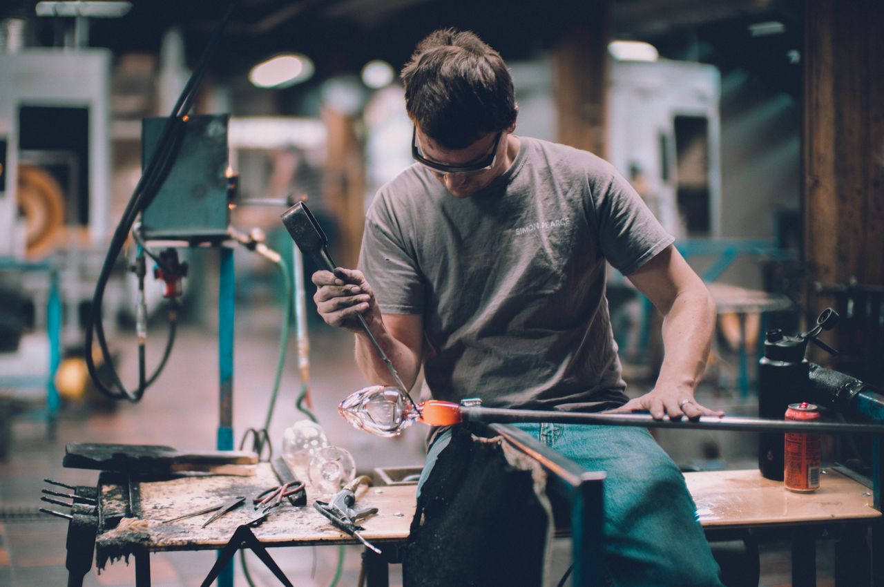 A glassblower molding red-hot glass using a long rod and a large, special knife. He is seated on a wooden bench, surrounded by tools.