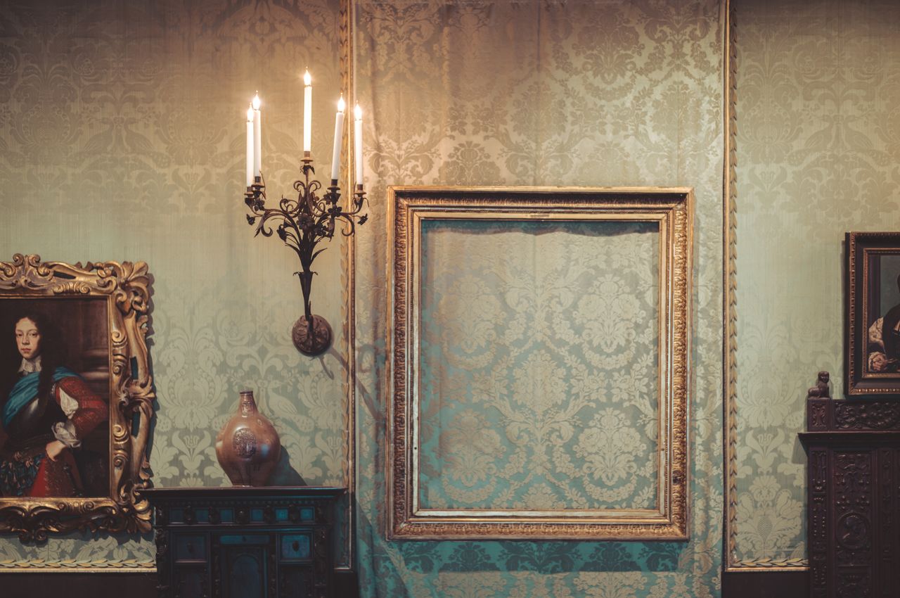 A a large, empty picture frame in a room with ornate wallpaper, candles, old paintings, and antique furniture.