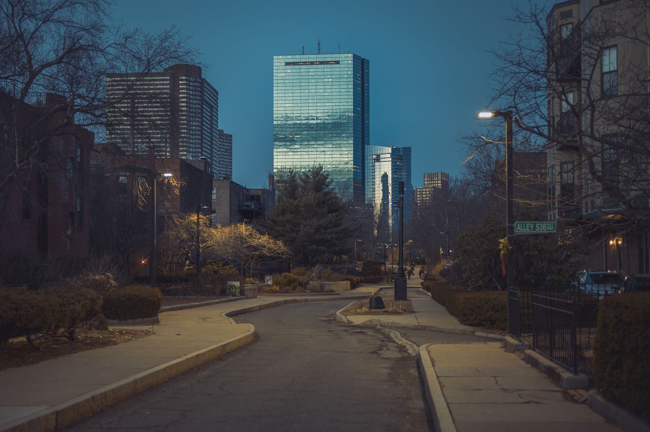 A skinny and bendy road in Boston, dark from dusk, leads the eye to Boston's largest skyscraper, whose mirror glass windows are reflecting the last sun.