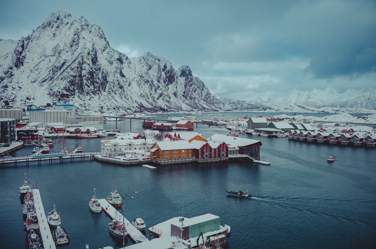 An aerial view of a harbor with traditional fishing boats docked on jetties and majestic snow-covered mountains providing a stunning backdrop.
