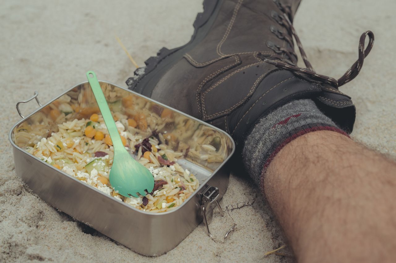 A meal in an aluminum container on the beach beside a hiking boot.