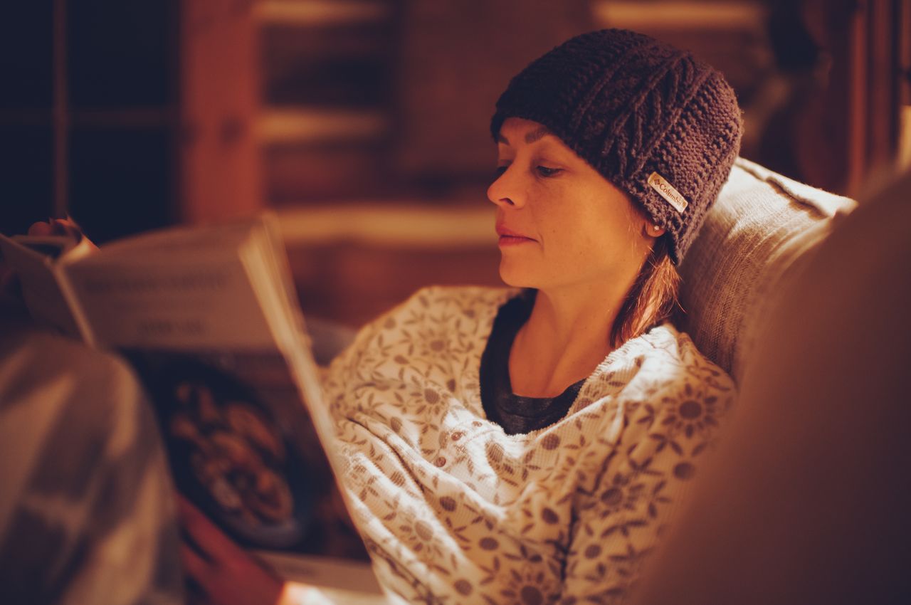 A woman, dressed in pajamas and a winter hat, is sitting on a sofa reading a book, surrounded by the softly lit warmth of a log cabin.