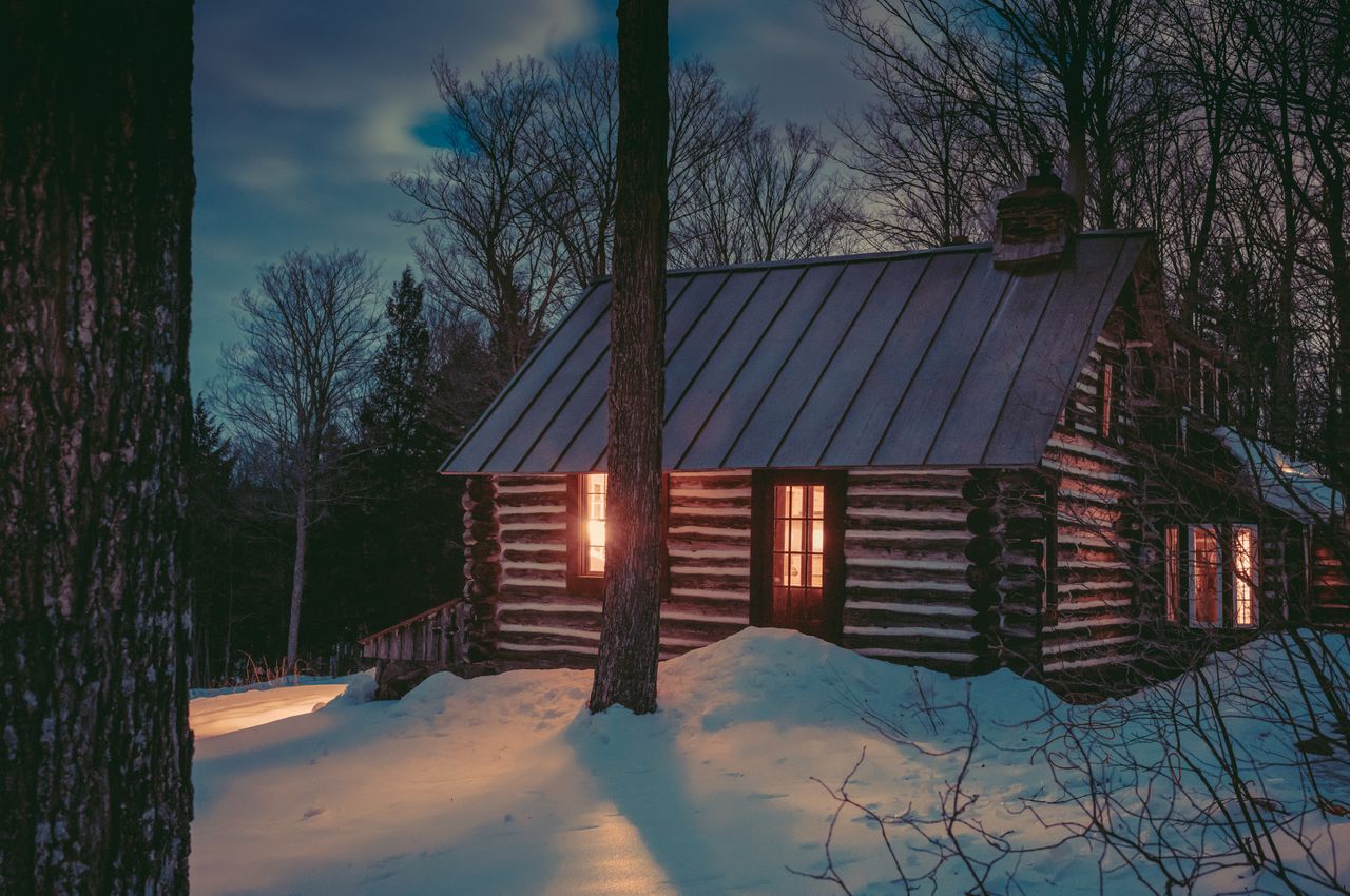 A log cabin nestled among trees, set in the evening's darkness, its interior lights reflecting on the snow and creating a peaceful atmosphere.