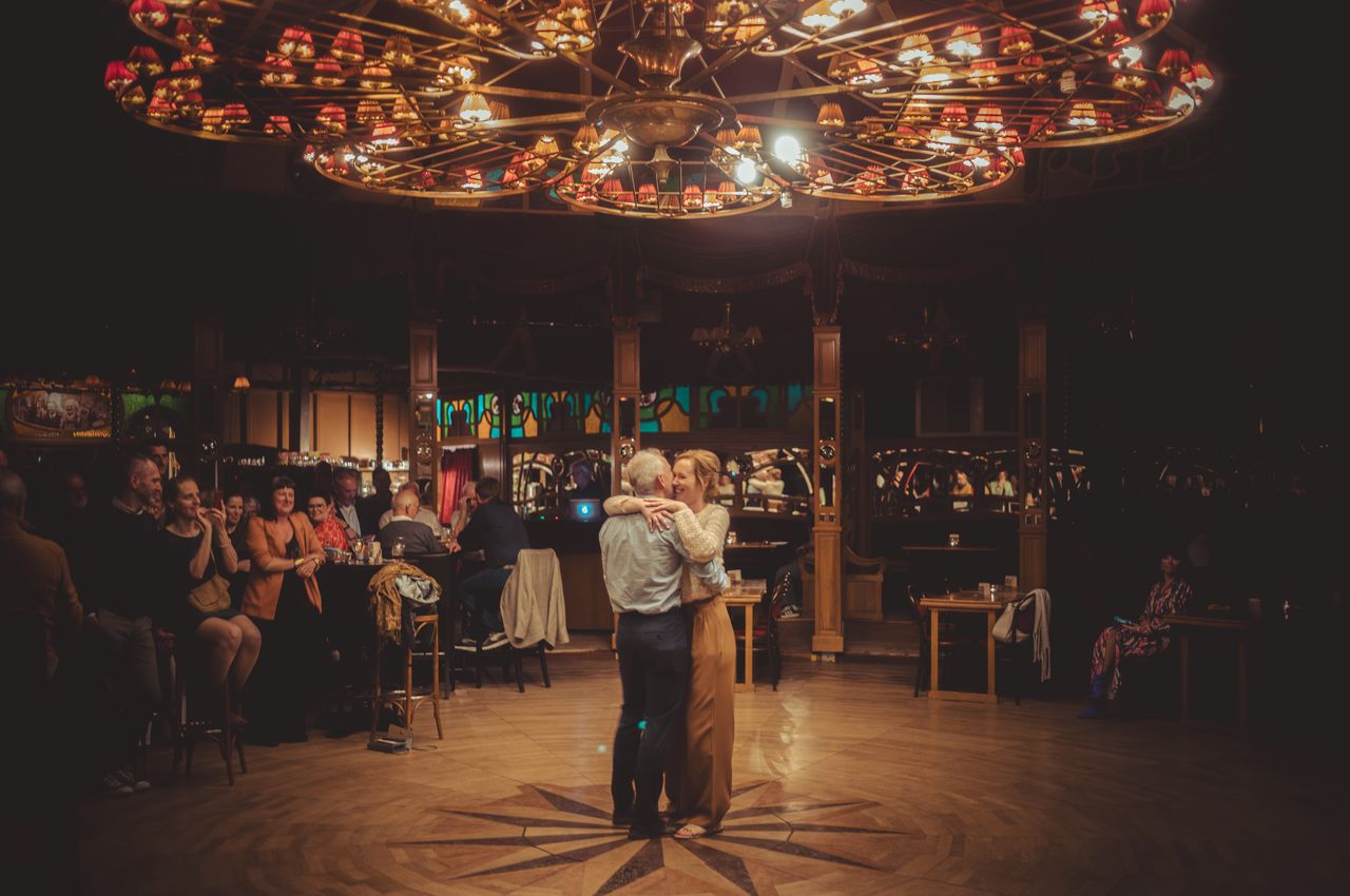 A couple dances beneath a grand chandelier, surrounded by family and friends.