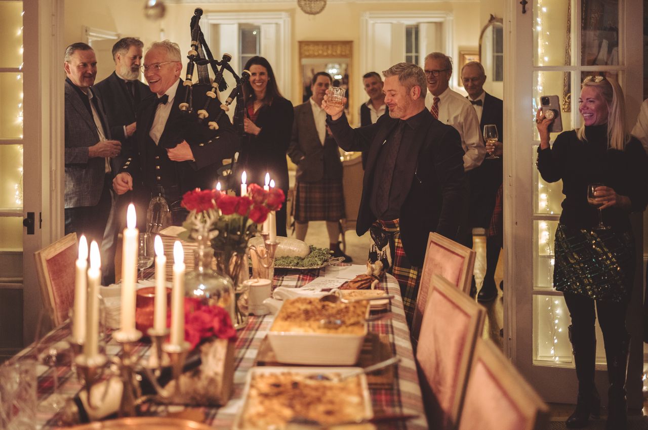 People in Scottish kilts gather around a well-stocked table in a candlelit room, toasting together after enjoying a bagpiper's performance.