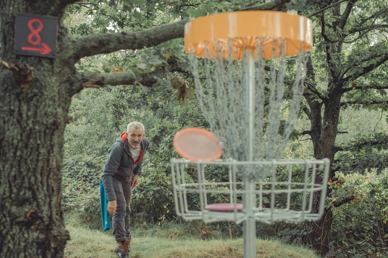 A man throws a disc toward a disc golf basket near a tree marked with hole 8.