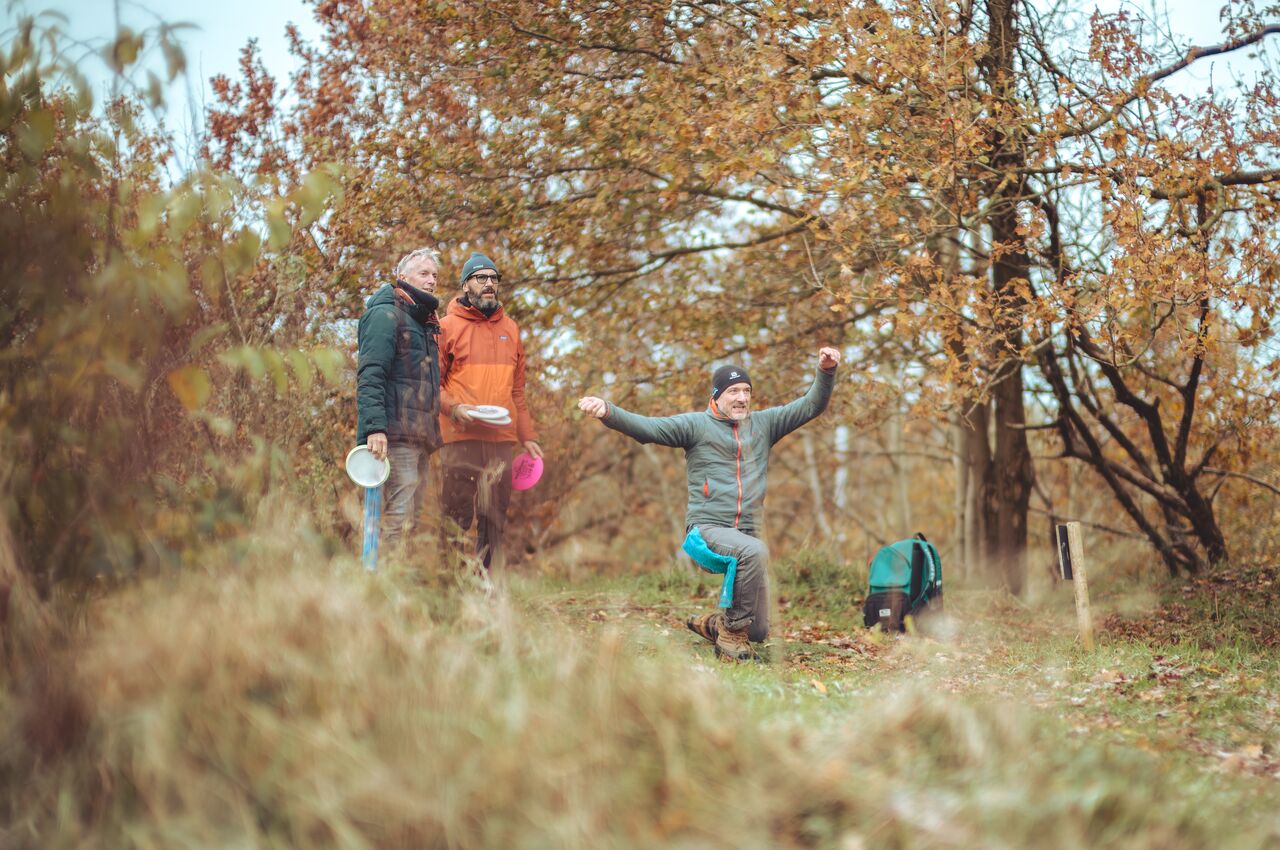 Three people outdoors in autumn, playing disc golf, one cheering.