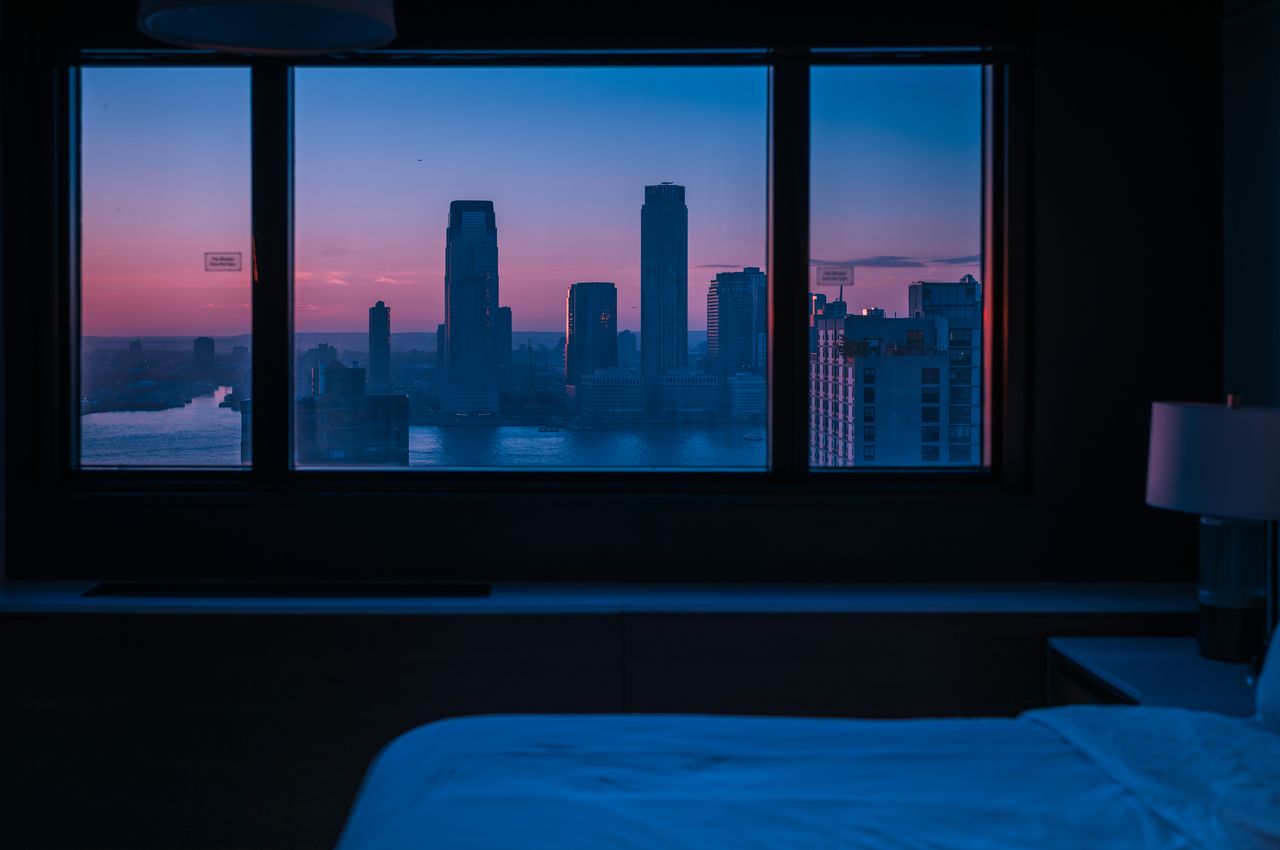 New York City's skyline seen through a window from a dimly lit hotel room.