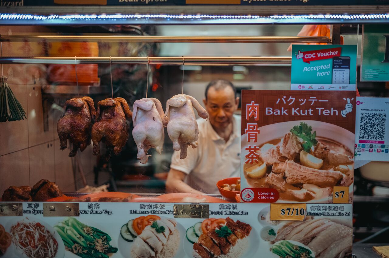 A food stall with chickens hanging behind a glass window, and a chef working in the background.