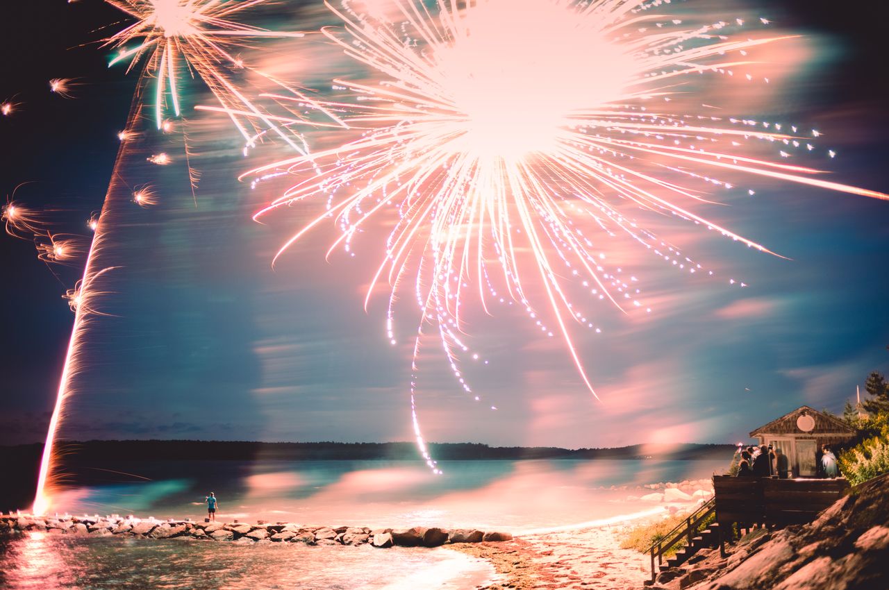 Fireworks explode over the ocean, ignited by a person standing on a jetty that extends into the water.