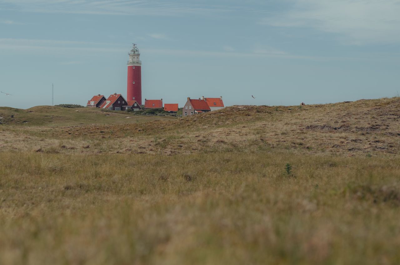 A red lighthouse and red-roofed houses viewed from a distance across grassy dunes.