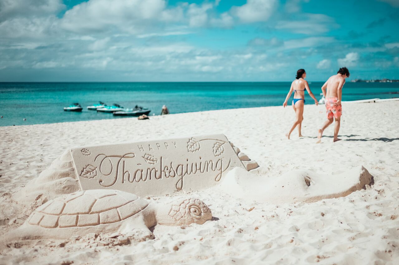 Sand sculpture reading 'Happy Thanksgiving' on a beach, with a couple walking nearby on the beach.