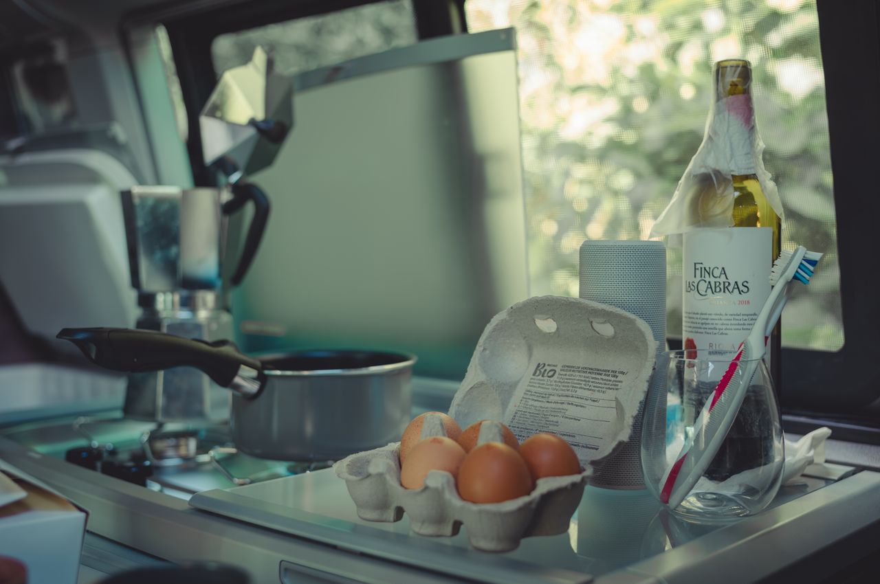 Interior of a small camper van with a Moka pot brewing coffee, a half-empty bottle of red wine, toothbrushes in a glass, eggs in a carton, and a portable speaker.