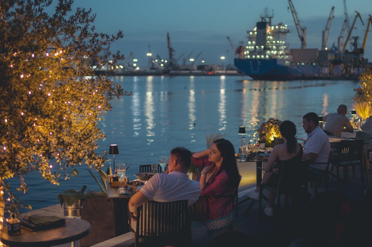 People dining outdoors with a view of the illuminated harbor and ships.