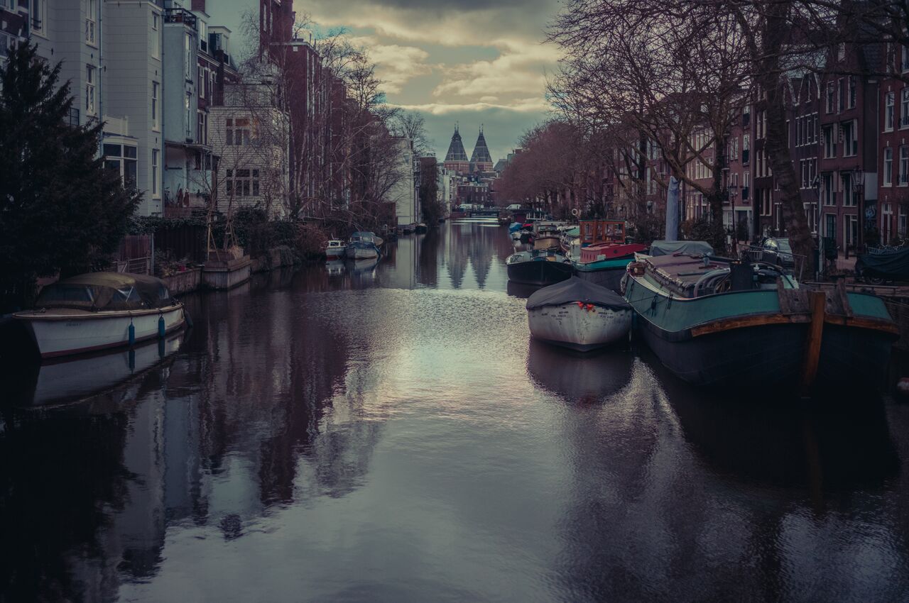 Amsterdam canal with houseboats, calm water reflections, and the Rijksmuseum in the distance under an overcast sky.