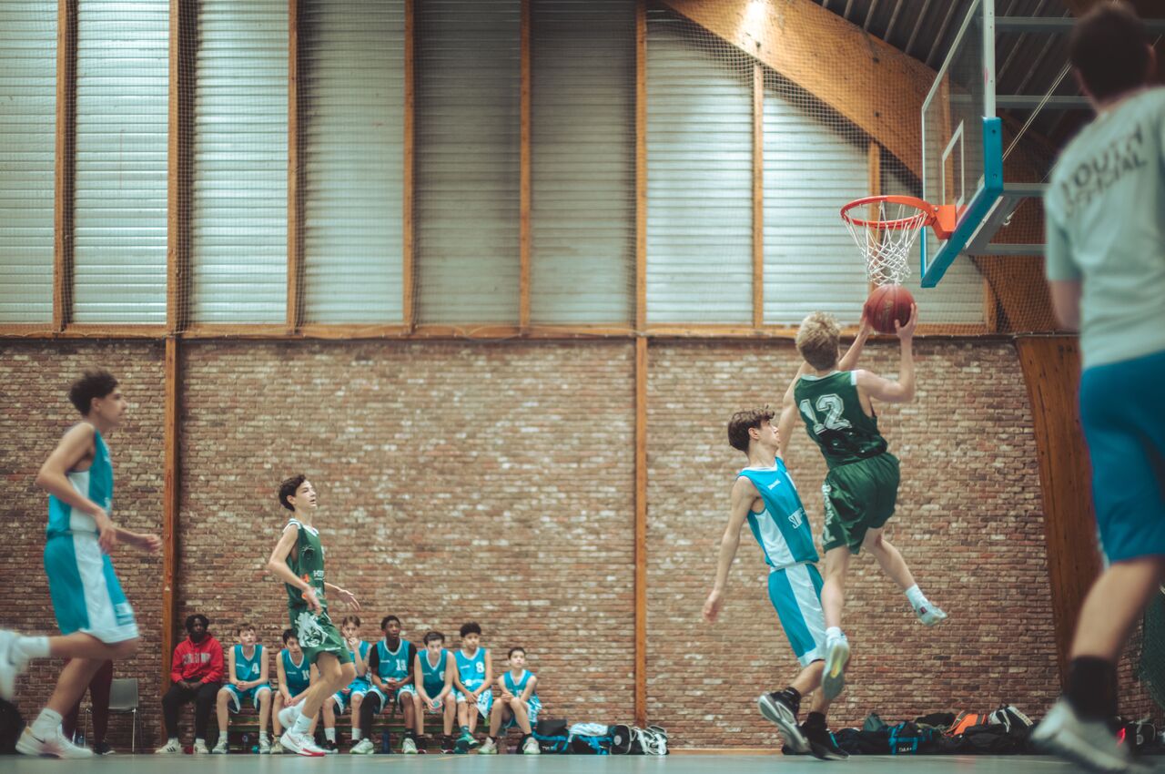 A basketball player in a green jersey jumps for a layup while a defender in blue attempts to block his shot.