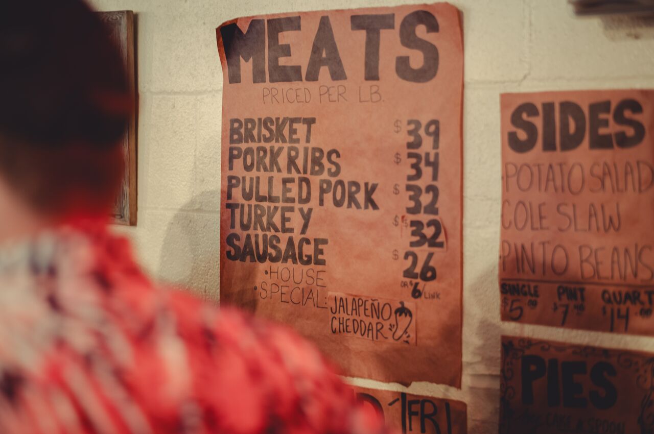 Handwritten barbecue menu on butcher paper hanging on a wall. It lists meats and sides with prices per pound, including brisket, pork ribs, pulled pork, turkey, sausage, and jalapeño cheddar sausage.