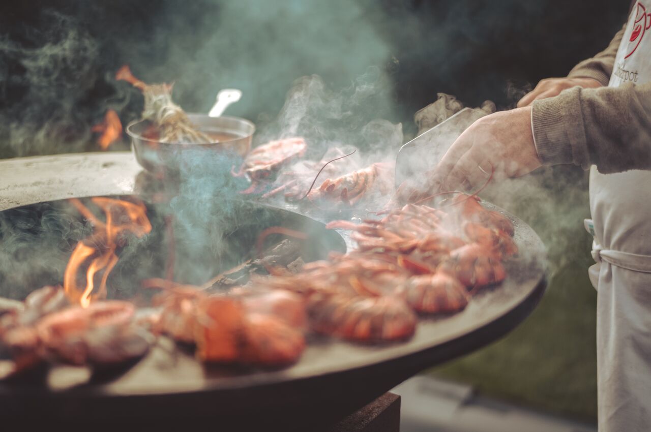 Lobsters are grilling on a round steel plate above an open flame, with a person flipping them using tongs.