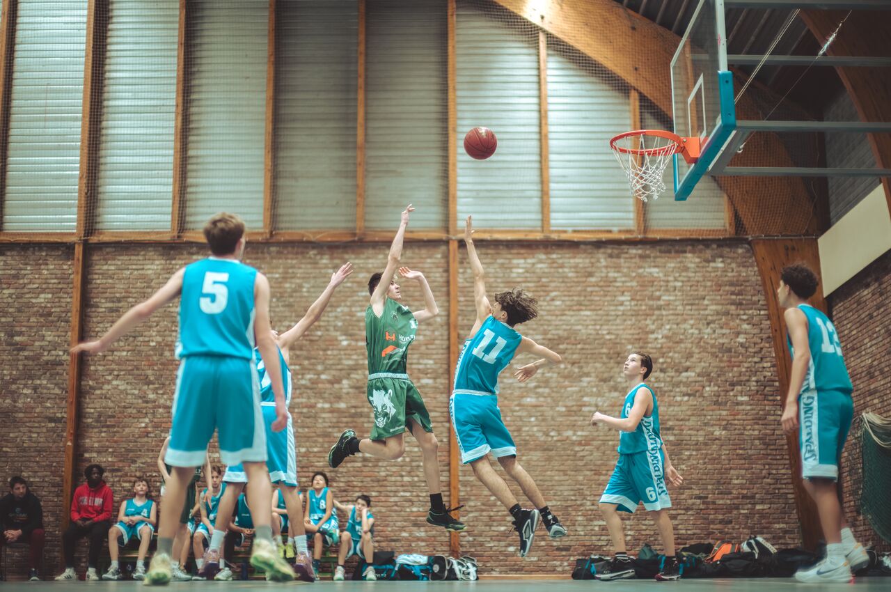 A basketball player in a green jersey jumps to take a shot while a defender in blue attempts to block his shot.