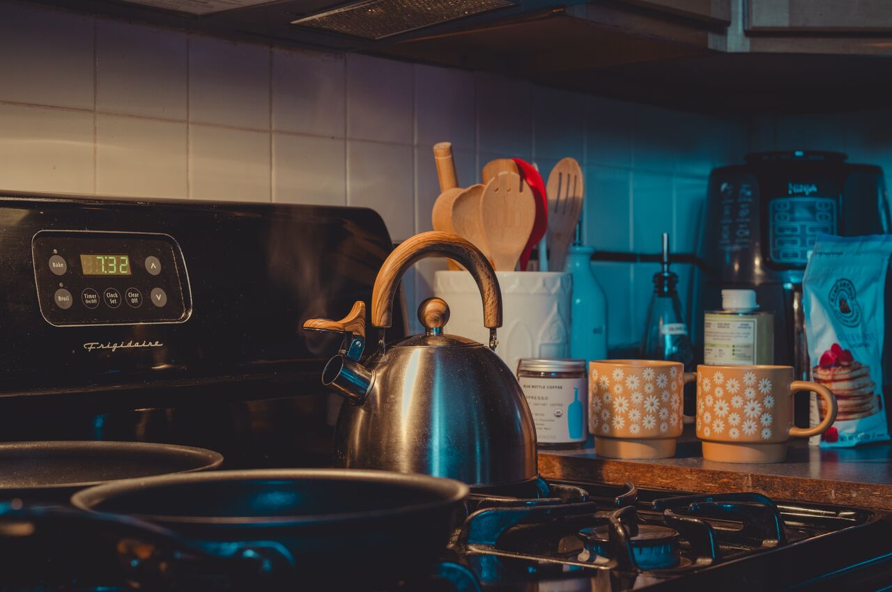 A steaming kettle on a gas stove, with coffee mugs and pancake mix in the background.