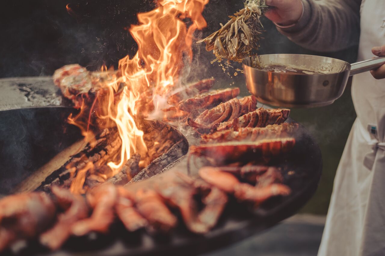 Lobsters grilling over open flames as someone applies metled butter using a bundle of herbs.