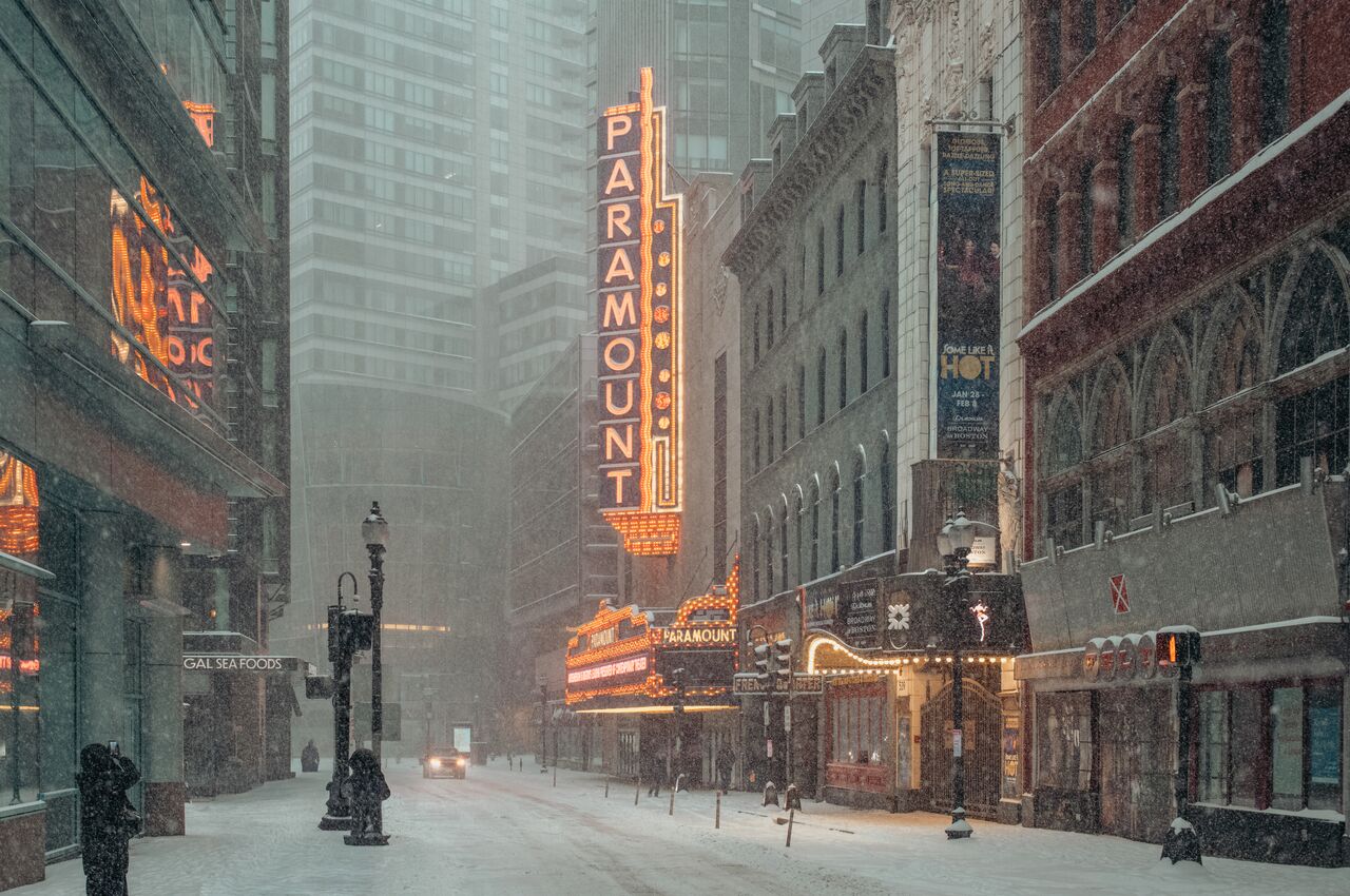 An empty street in downtown Boston during a snowstorm, lit by the orange Paramount Theatre sign.