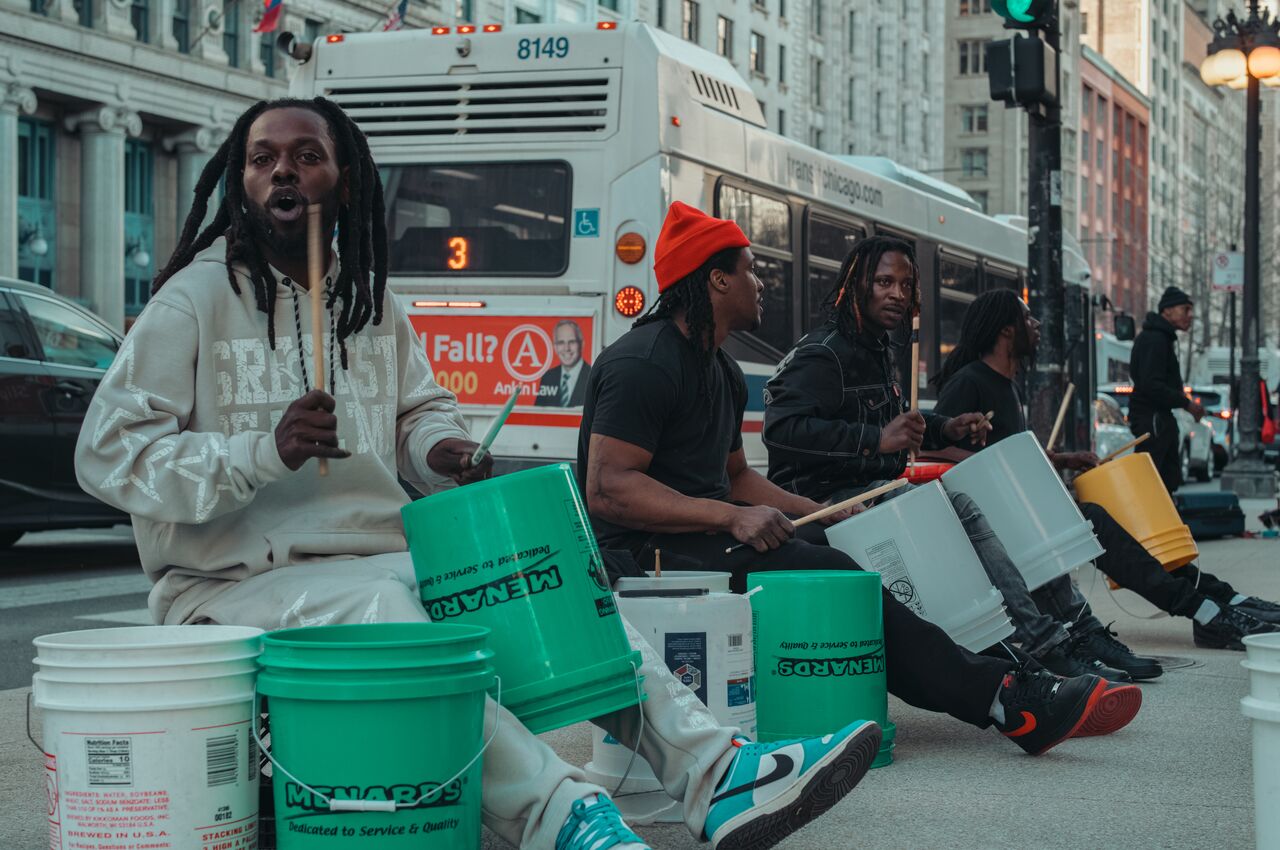 Four men sit on a sidewalk in downtown Chicago and play drums on cheap plastic buckets.