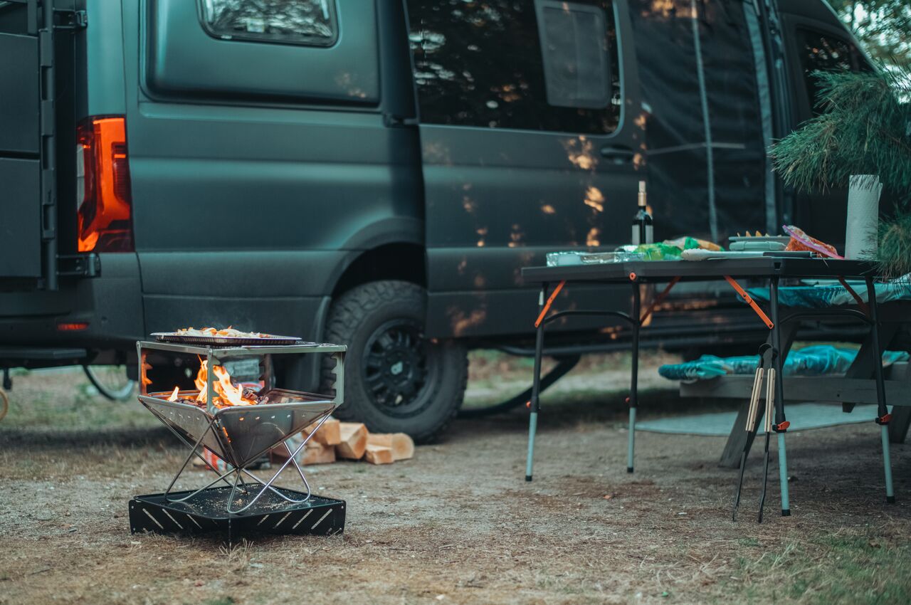 A small fire burns in a portable grill beside a camping table and a camper van.