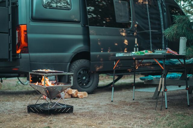 A small fire burns in a portable grill beside a camping table and a camper van.
