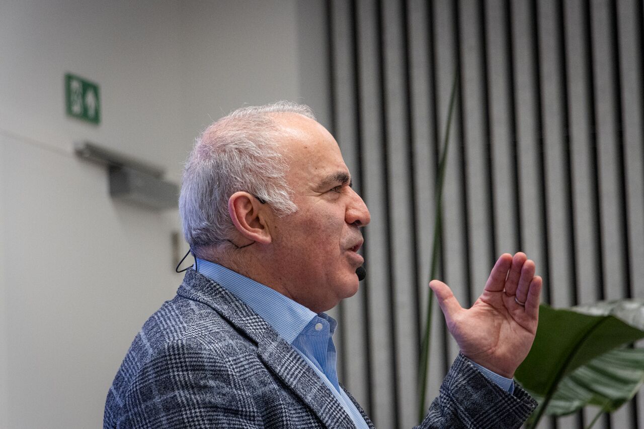 Garry Kasparov in profile, wearing a checked blazer, raising one hand as he speaks in front of a dark slatted wall.