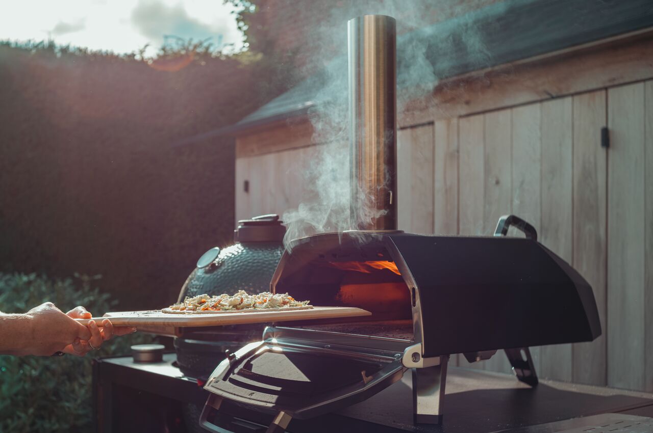 A homemade pizza is being slid into a smoking outdoor pizza oven.