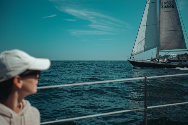 A person in a white cap and sunglasses watches a sailboat gliding by.
