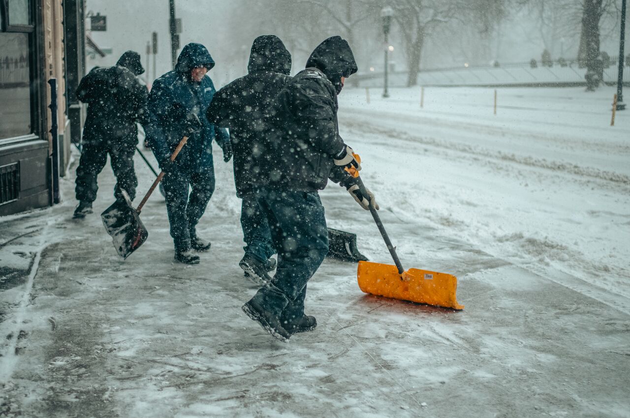 Four workers in hooded jackets shovel snow along the curb.