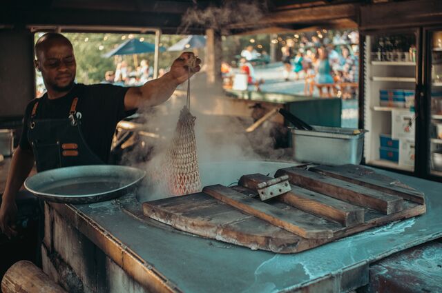 A cook lifts a mesh bag of freshly steamed lobster from a steaming kettle.