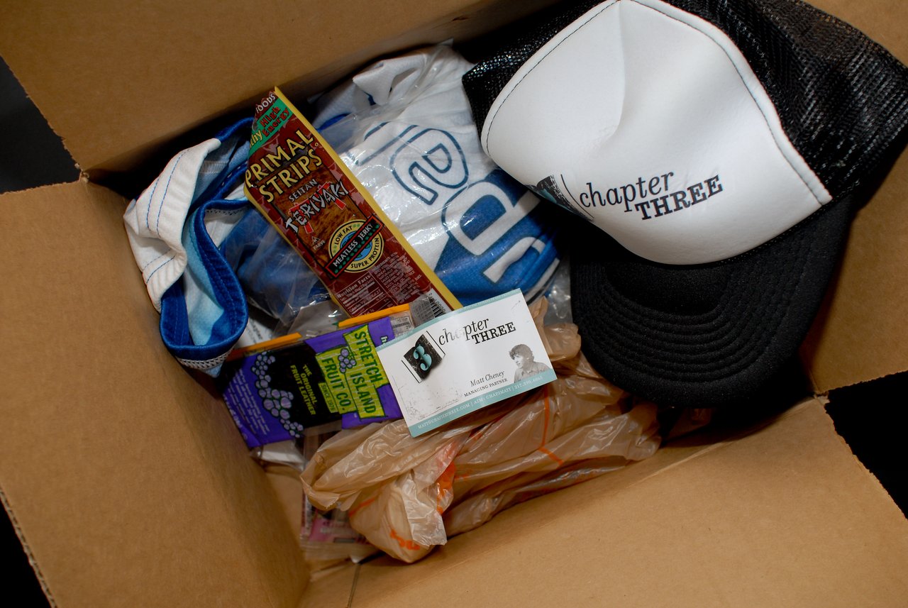 An open box contains a hat, a Drupal jersey, snacks, a business card, and other small items.